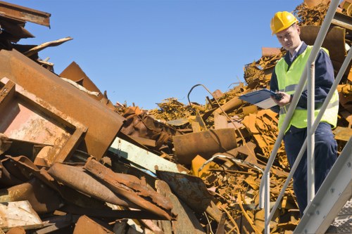 A construction or waste management inspector, wearing a yellow safety helmet and high-visibility yellow vest over a dark blue jacket, is standing on a metal staircase next to a large pile of rusted metal debris. The debris consists of various sheets, beams, and other metal objects with a heavily corroded, reddish-brown finish, scattered in a chaotic manner. The inspector is holding a clipboard and appears to be reviewing or recording details about the waste. The environment is outdoors with clear blue sky overhead, suggesting a bright, sunny day. The scene is set on a site that could be part of a rubbish disposal or recycling area, relevant to businesses offering waste removal services in Hackney or nearby areas. Business Waste Removal Hackney operates in this context, supporting efficient clearance and disposal of such construction or industrial waste materials.