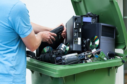 A person wearing a light blue t-shirt is shown loading electronic waste, including a black desktop computer tower with an open side panel and visible internal components, onto a green waste skip or bin. The electronic waste collection contains various items such as CDs, a digital camera, and other electronic devices and accessories, all cluttered together loosely inside the bin. The scene is set outdoors, likely on a driveway or paved area, with the focus on the bin and the person's hands. The image documents the disposal of obsolete electronics, relevant to waste collection and recycling services available in Hackney, as provided by Business Waste Removal Hackney. The environment appears clean and well-maintained, with natural daylight illuminating the scene, emphasizing the variety of materials and textures, such as the plastic casing of the electronics and the smooth surface of the green bin, conveying the process of electronic waste disposal clearly.