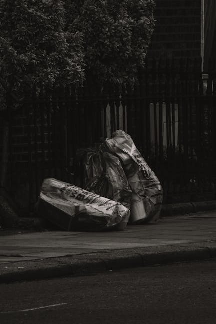 Three large black garbage bags made of thick, crinkled plastic are placed on the pavement near a black metal fence and a wooden fence, with a green leafy tree visible in the background. The bags appea
