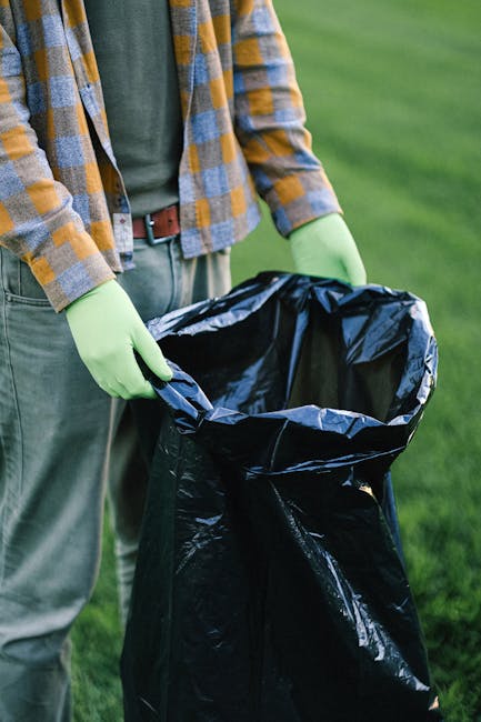 A person dressed in orange work overalls and white shoes is standing on a plain, light grey floor with a neutral, light background. They are holding two large blue plastic rubbish bags, each filled wi