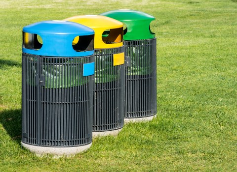 Three cylindrical outdoor rubbish bins with slatted black plastic bodies are positioned on a well-maintained grassy area adjacent to a concrete pavement. Each bin has a vibrant, rounded lid in yellow, green, and blue respectively, with openings for waste disposal and small rectangular blue or yellow labels. The lids appear smooth and slightly glossy, contrasting with the textured, matte finish of the slatted bodies. The bins are situated in a line, with the blue-lidded bin on the left, the yellow-lidded in the center, and the green-lidded on the right. They are set against a background of lush, green grass, suggesting a park or communal outdoor space that could be typical of areas in Hackney or similar London boroughs. The lighting is natural, likely midday sunlight, casting soft shadows on the ground and emphasizing the vivid colors of the lids. This arrangement is indicative of waste management infrastructure employed by local authorities or waste removal services such as Business Waste Removal Hackney to facilitate rubbish collection and recycling efforts within urban or semi-urban environments.